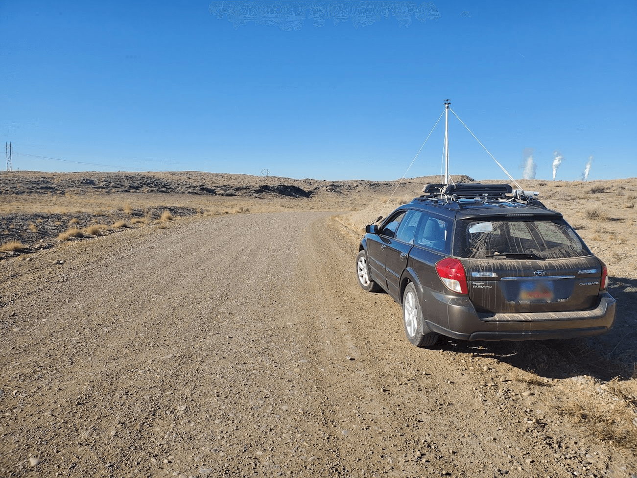 A hatchback car is parked on the side of a dirt road adjacent to short grasses. The roof of the car is equipped with environmental monitoring equipment, including a TriSonica<sup>®</sup> Mini Wind and Weather Sensor and an air intake for a trace gas analyzer. An industrial facility or oil refinery is visible on the distant horizon.