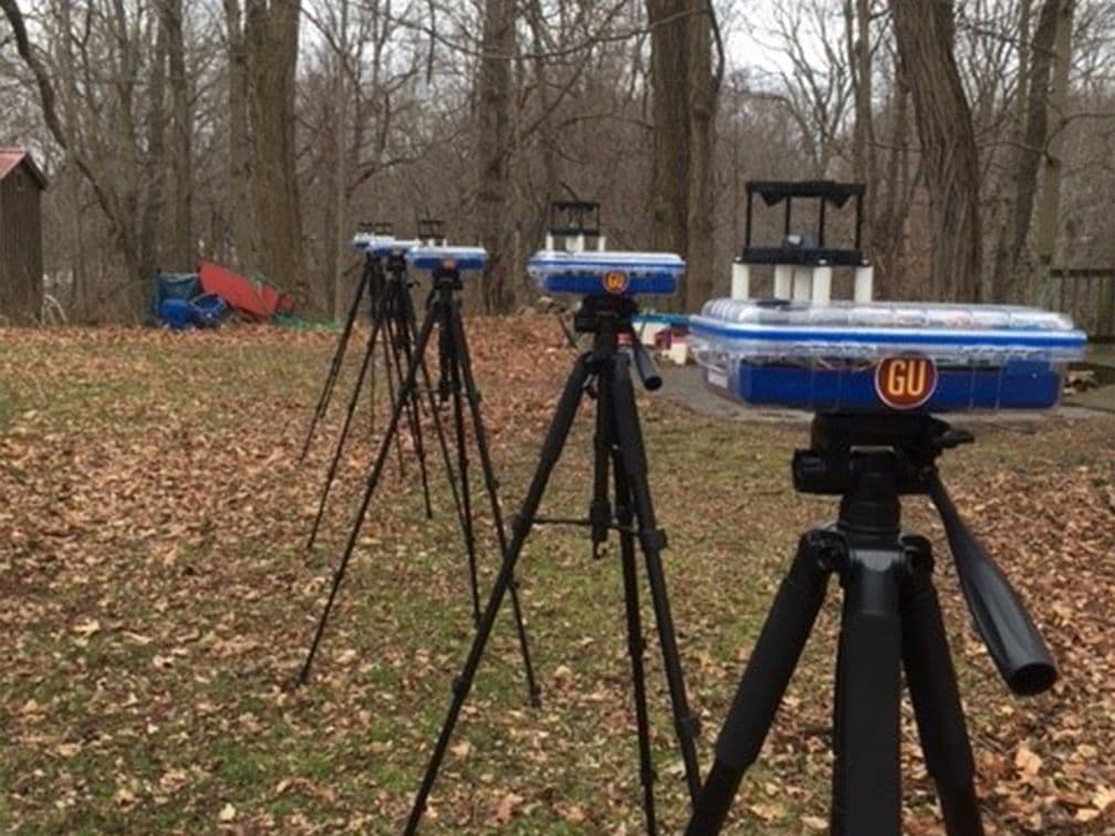 A row of five small tripods support five identical sensor platforms, each featuring a LI-550F TriSonica Mini Wind and Weather Sensor. A leafless woodland canopy stands in the background, while sparse leaf litter covers the mowed grass in the foreground.